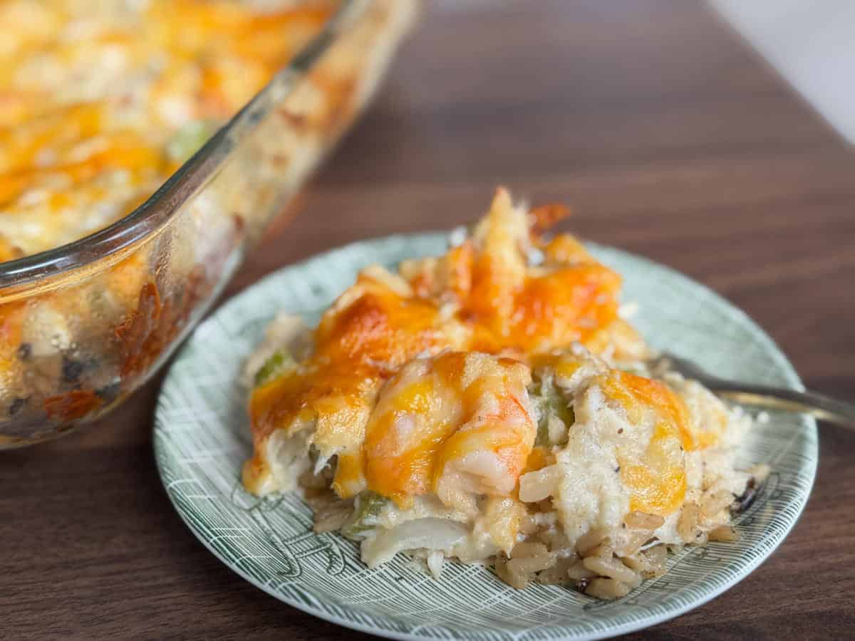 A serving of cheesy casserole with shrimp, vegetables, and rice sits on a green patterned plate next to a larger glass baking dish on a wooden table.