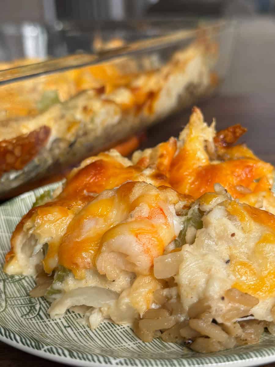A close-up of a plate with a serving of cheesy rice casserole containing shrimp and vegetables, with the casserole dish visible in the background.
