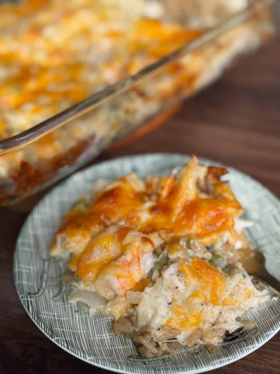 A close-up of a plate with a serving of cheesy casserole containing rice, shrimp, and vegetables. A glass baking dish with more of the casserole is visible in the background on a wooden surface.