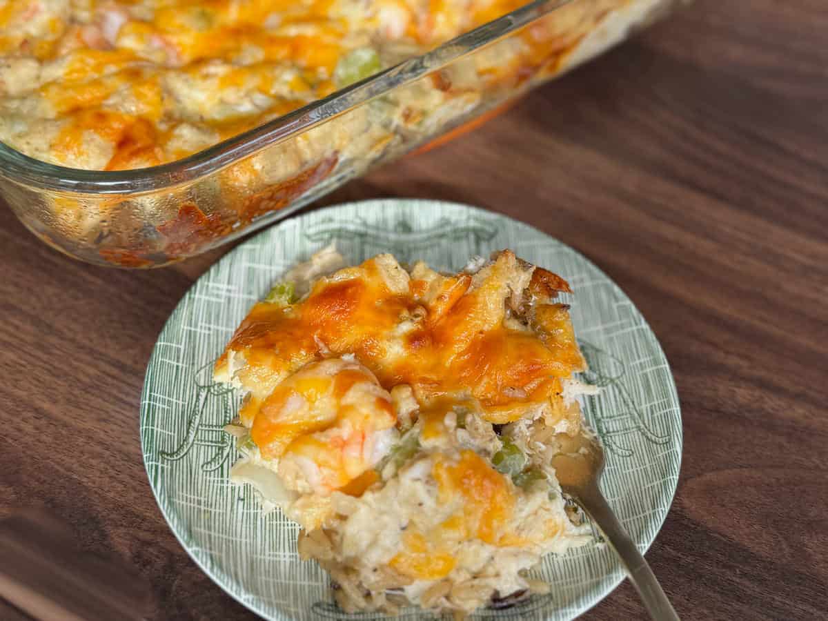 A serving of cheesy casserole with visible shrimp and vegetables sits on a green patterned plate, next to a glass baking dish containing the rest of the casserole, all on a wooden surface.