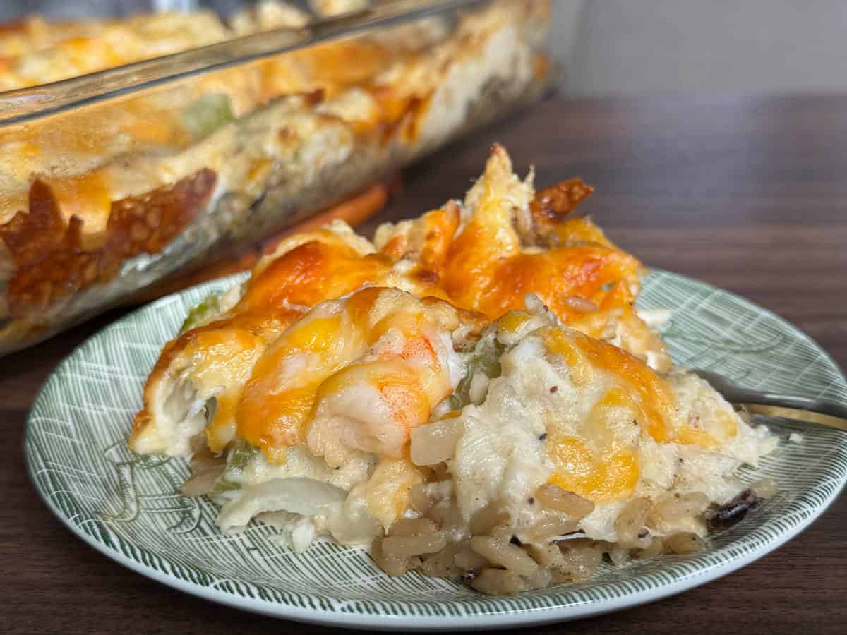 A plate with a serving of cheesy seafood and rice casserole, showing shrimp, rice, and vegetables, sits in front of a glass baking dish with more casserole on a wooden surface.