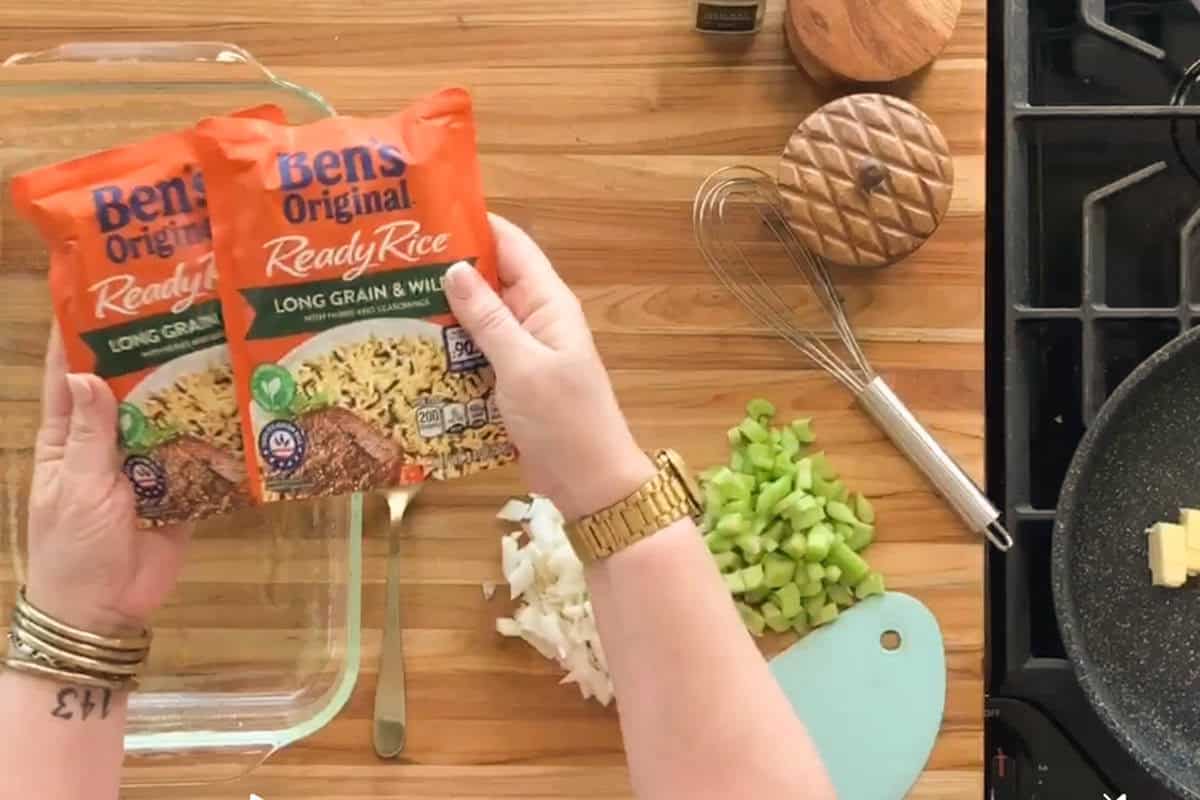 A person holding two packets of Ben’s Original Ready Rice over a glass baking dish. Chopped onions and celery are on a cutting board, and a pan with butter is on a stove to the right. Cooking utensils are visible on the counter.