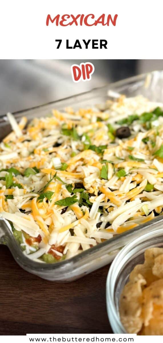 A glass dish filled with Mexican 7 layer dip, topped with shredded cheese, sliced olives, and green onions sits next to a bowl of tortilla chips on a wooden surface. Text on the image reads “MEXICAN 7 LAYER DIP.”.