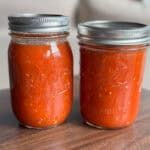 Two glass mason jars filled with homemade tomato sauce sit side by side on a wooden surface. Both jars are sealed with metal lids and contain a thick, chunky red sauce.