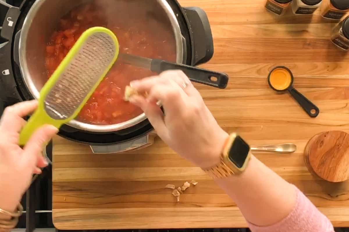 A person grates a piece of ginger over a pot of tomato sauce on a wooden countertop. A measuring spoon, small container, and spice jars are arranged nearby.