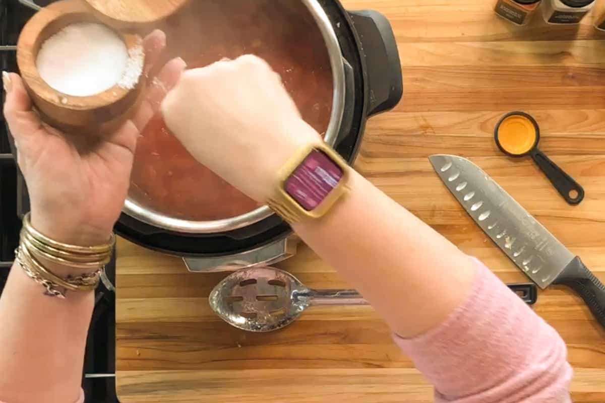A person wearing a gold wristwatch adds salt to a pot of red sauce on a stove. A knife, slotted spoon, and measuring spoon are on the wooden counter nearby.