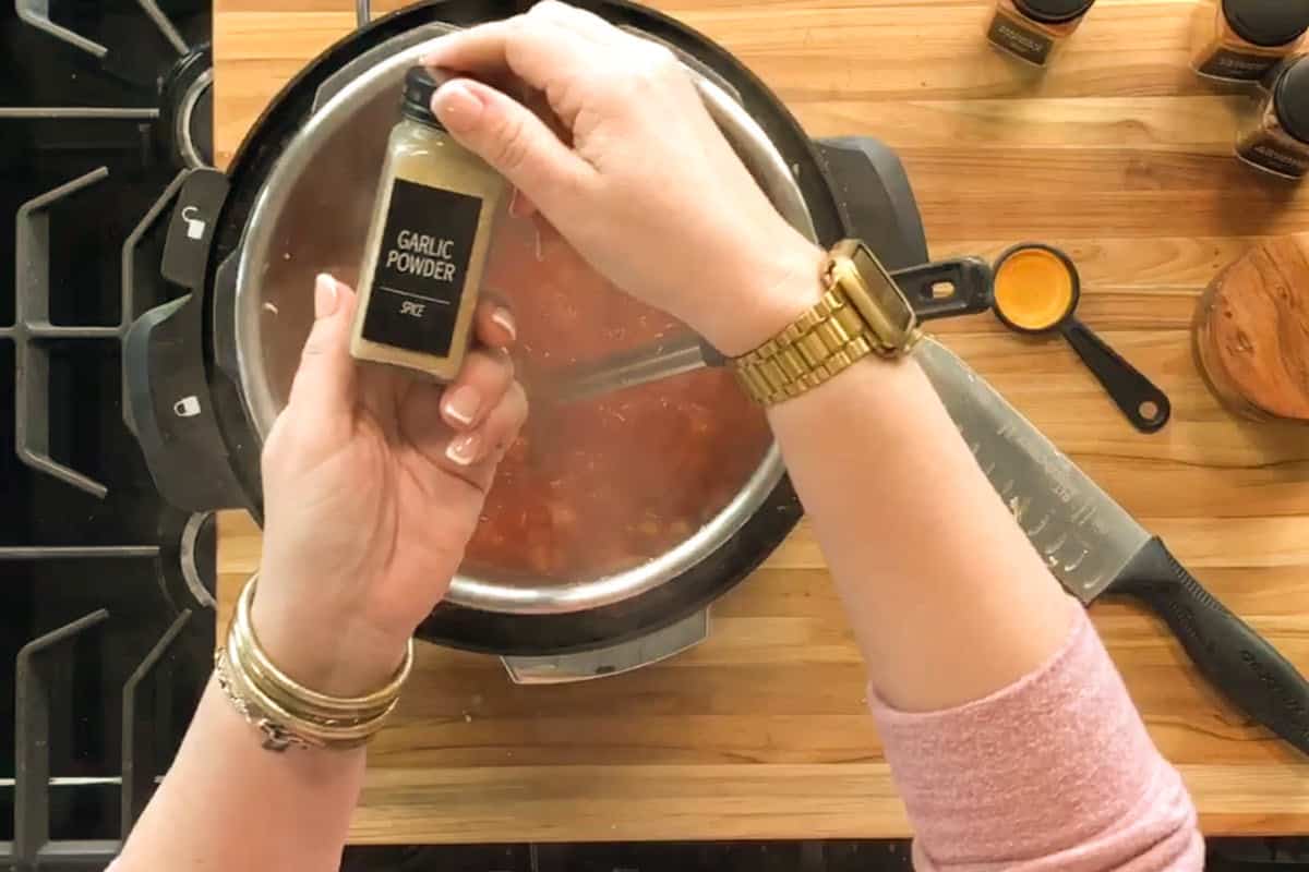 A person holding a bottle labeled "Garlic Powder" above a pot of food on a stovetop, with a cutting board, measuring spoon, and knife visible on the wooden counter.