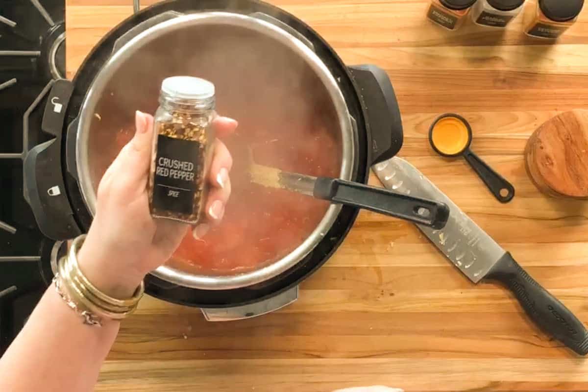 A hand holds a small jar labeled "Crushed Red Pepper" above a pot of simmering tomato sauce on a wooden countertop, with a knife, spoon, and other spices nearby.