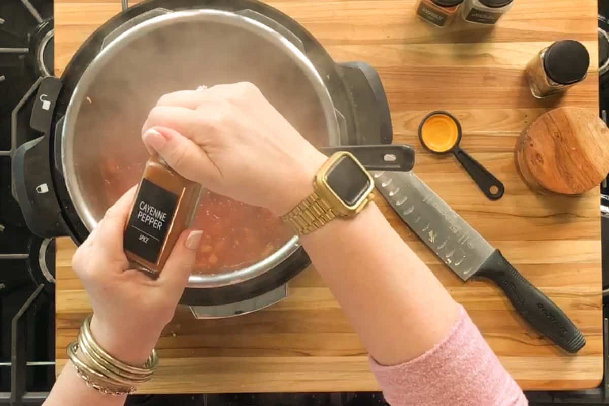 A person holding a cayenne pepper spice bottle over a steaming pot on a wooden cutting board, surrounded by a knife, measuring spoons, and other spice jars. The person is wearing a gold watch and bracelets.