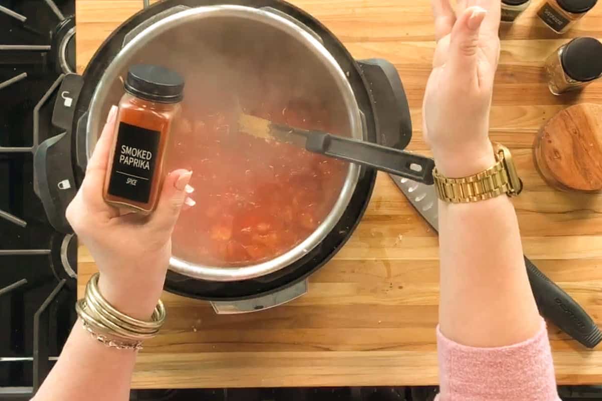 A person adds ingredients to a steaming pot on a stove while holding a jar labeled "smoked paprika." Kitchen utensils and spice jars are on a wooden countertop.