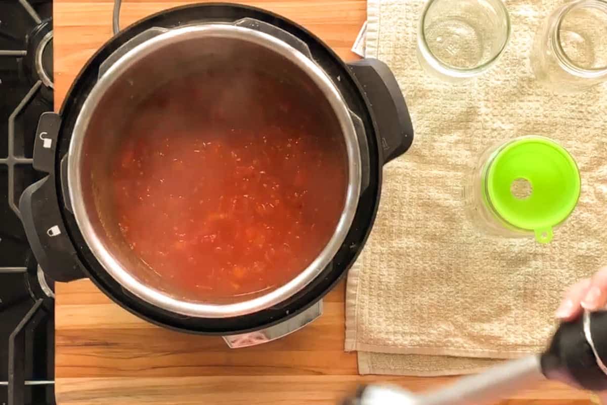 A pot of tomato sauce cooking on a stovetop next to empty glass jars on a towel, along with a green funnel and an immersion blender being held by a hand.