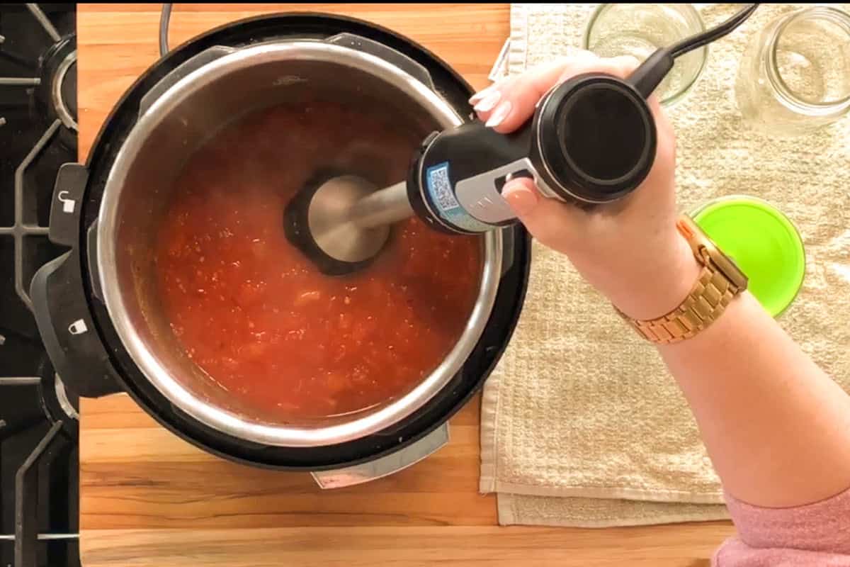 A person uses an immersion blender to blend tomato sauce inside a pressure cooker on a wooden countertop. Nearby are empty glass jars, a green lid, and a beige towel.