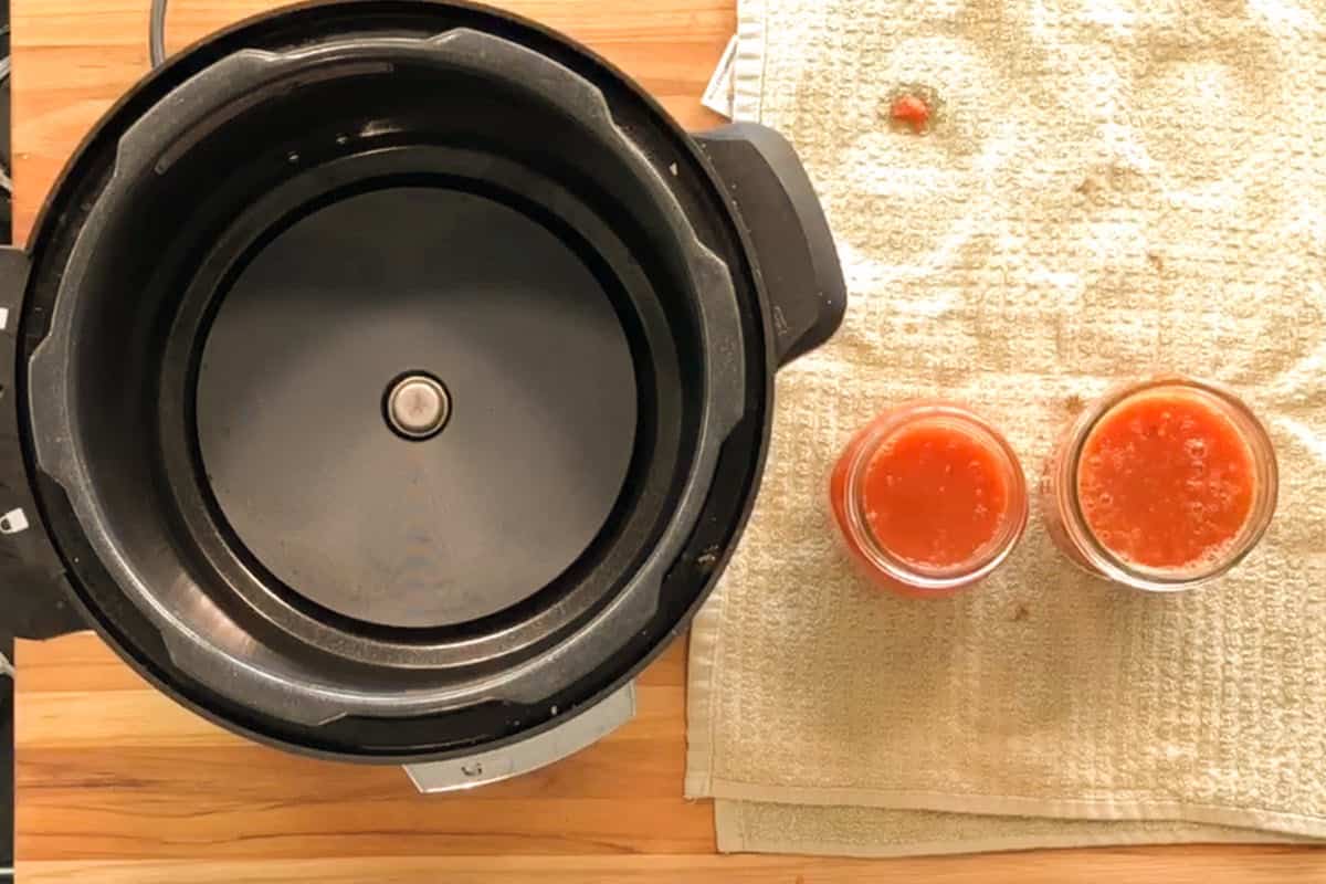 An open electric pressure cooker sits on a wooden surface next to a beige towel with two small glass jars filled with tomato sauce and a few sauce splatters on the towel.