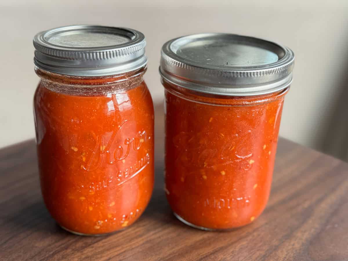 Two glass jars filled with homemade tomato sauce sit on a dark wooden surface. Both jars are sealed with metal lids and labeled with embossed brand names. The sauce inside is chunky and bright red.