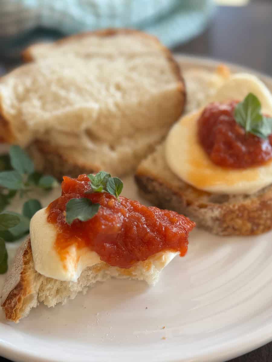 Two slices of bread topped with mozzarella cheese, tomato sauce, and fresh herbs are on a white plate. Additional bread slices and herbs are in the background.