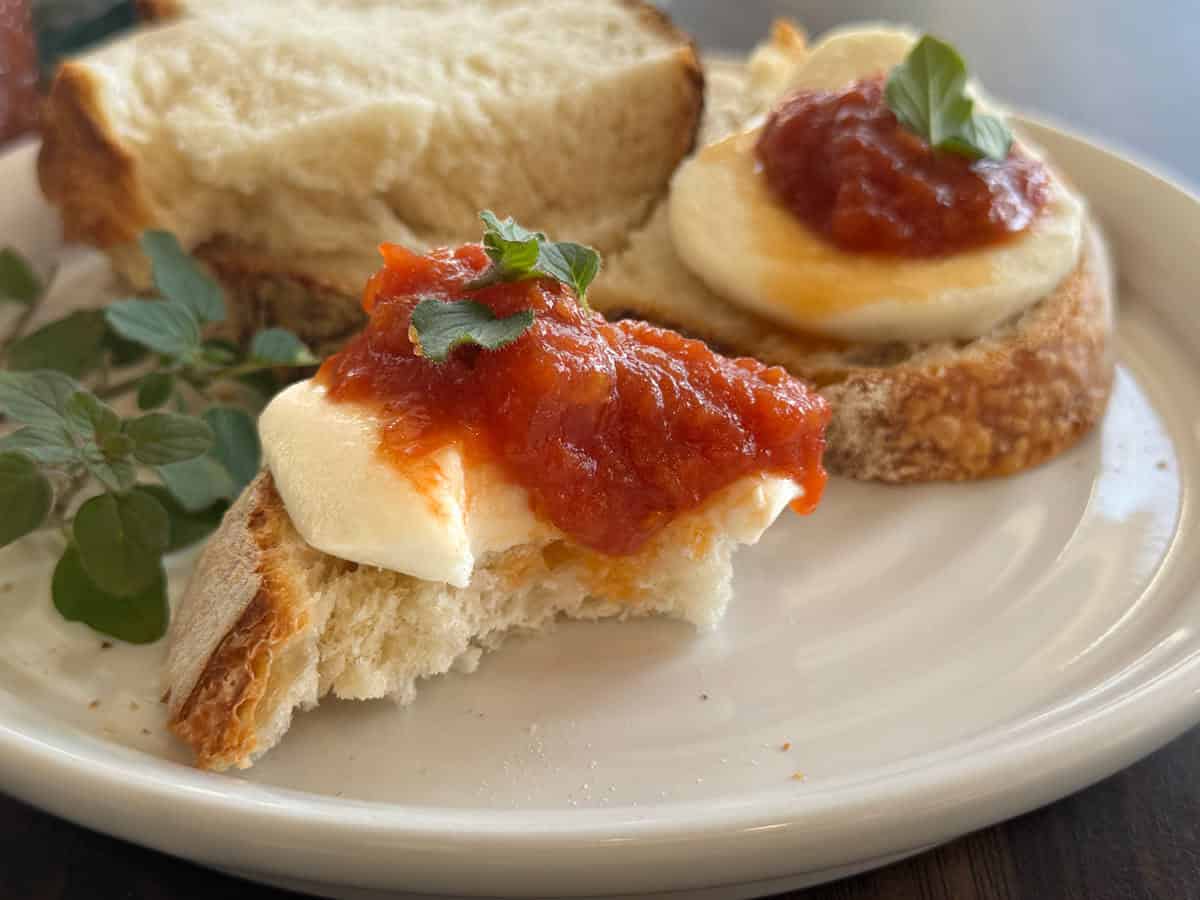 A close-up of a white plate with slices of bread topped with mozzarella cheese, tomato sauce, and fresh herbs. A sprig of green herbs is placed on the plate as garnish.