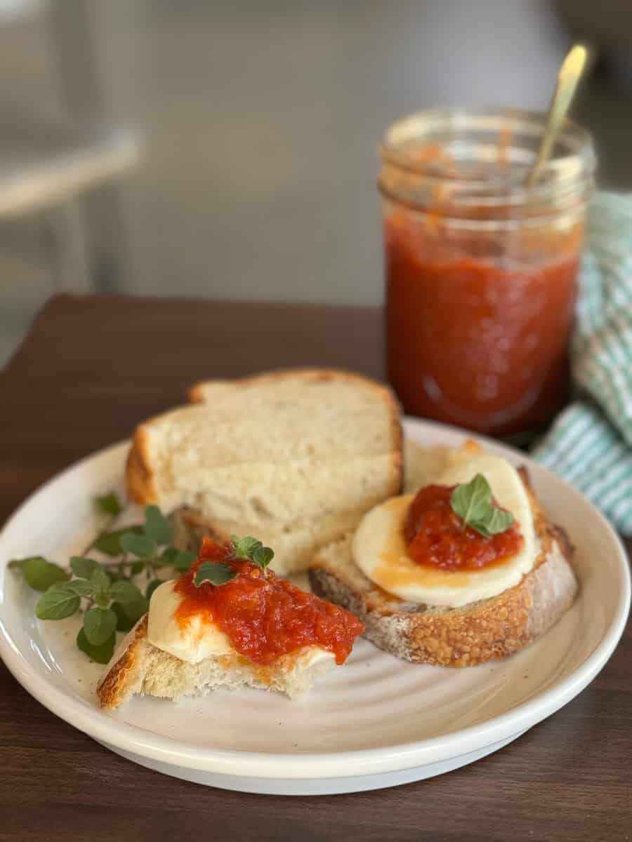 A plate with slices of bread, some topped with mozzarella, tomato sauce, and fresh oregano. A sprig of oregano and a jar of tomato sauce with a spoon are on the side, all set on a wooden table.