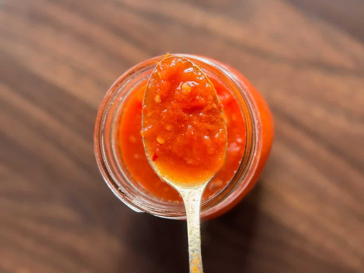 A close-up view of a spoonful of red sauce or salsa held over an open jar, with a wooden surface in the background.