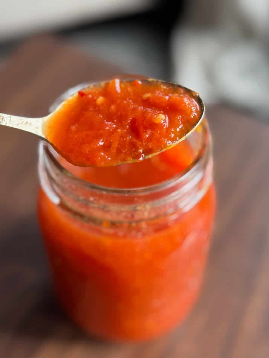 A close-up of a spoonful of chunky red tomato sauce held above a glass jar filled with the same sauce, on a wooden surface.