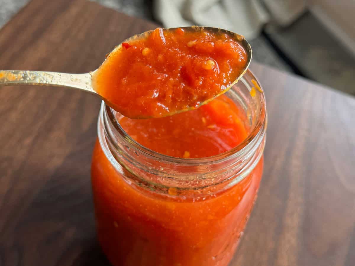 A metal spoon holding a portion of chunky red sauce hovers above an open glass jar filled with the same sauce, set on a wooden surface.