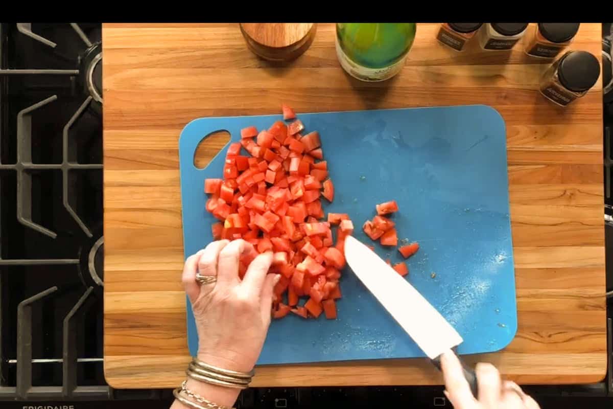 A person chops tomatoes into small cubes on a blue cutting board placed on a wooden countertop, with seasonings and a bottle nearby.