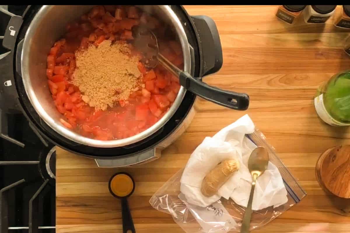 An Instant Pot on a stove contains chopped tomatoes and brown sugar being stirred with two utensils. Nearby, a piece of ginger sits in a plastic bag on a napkin, next to a measuring spoon and a green bottle.