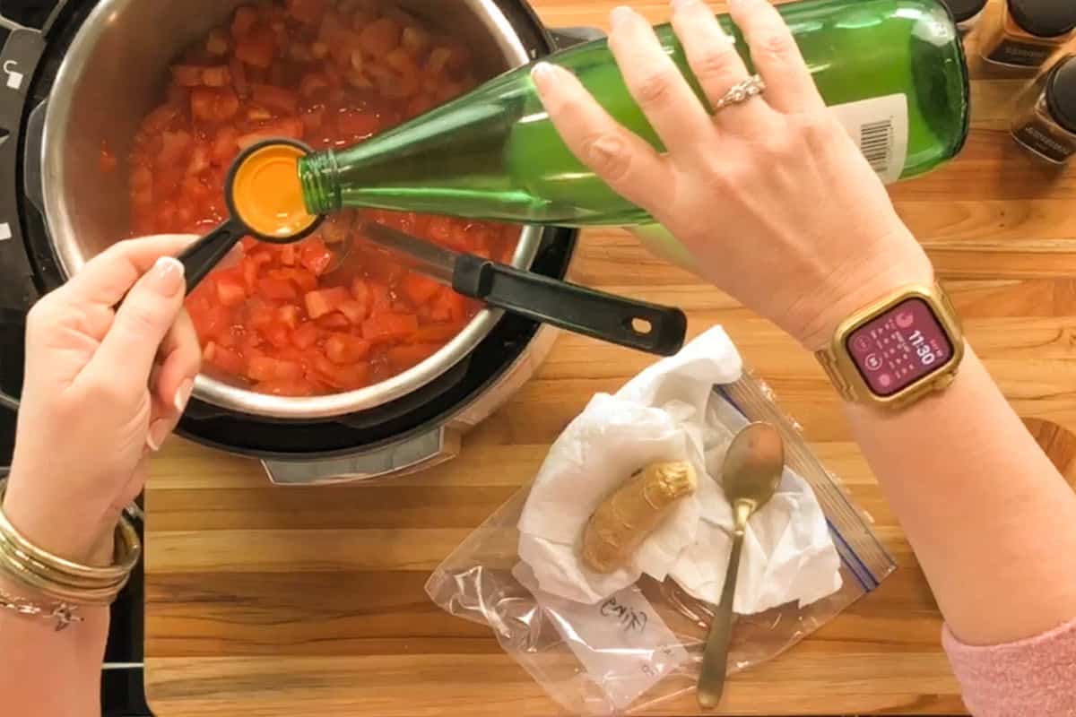 A person pours liquid from a green bottle into a measuring spoon over a pot of chopped tomatoes on a stove. A watch is on their wrist, and a ginger root with a spoon is beside the pot on a wooden counter.