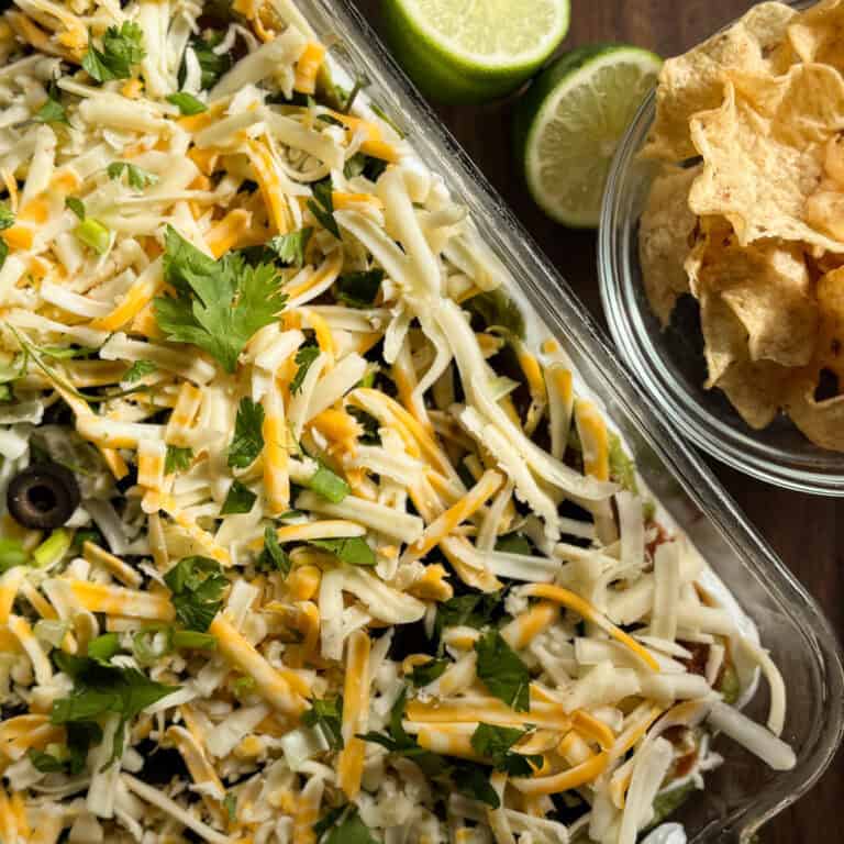 A close-up of a glass dish filled with shredded cheese, cilantro, and other toppings, next to a bowl of tortilla chips and halved limes on a wooden surface.