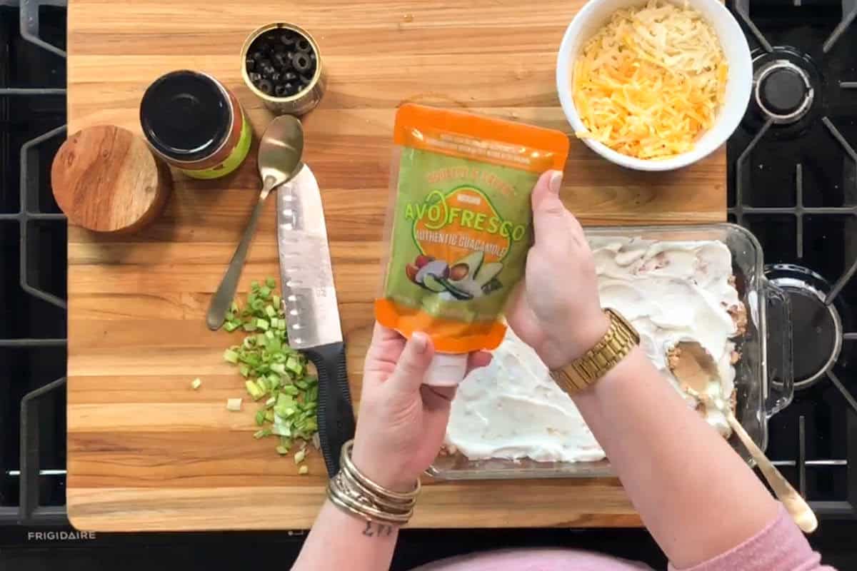 Person holding a pouch of avocado sauce above a casserole dish with a creamy white topping. Other ingredients, including chopped green onions, shredded cheese, olives, and spices, are on a wooden cutting board.