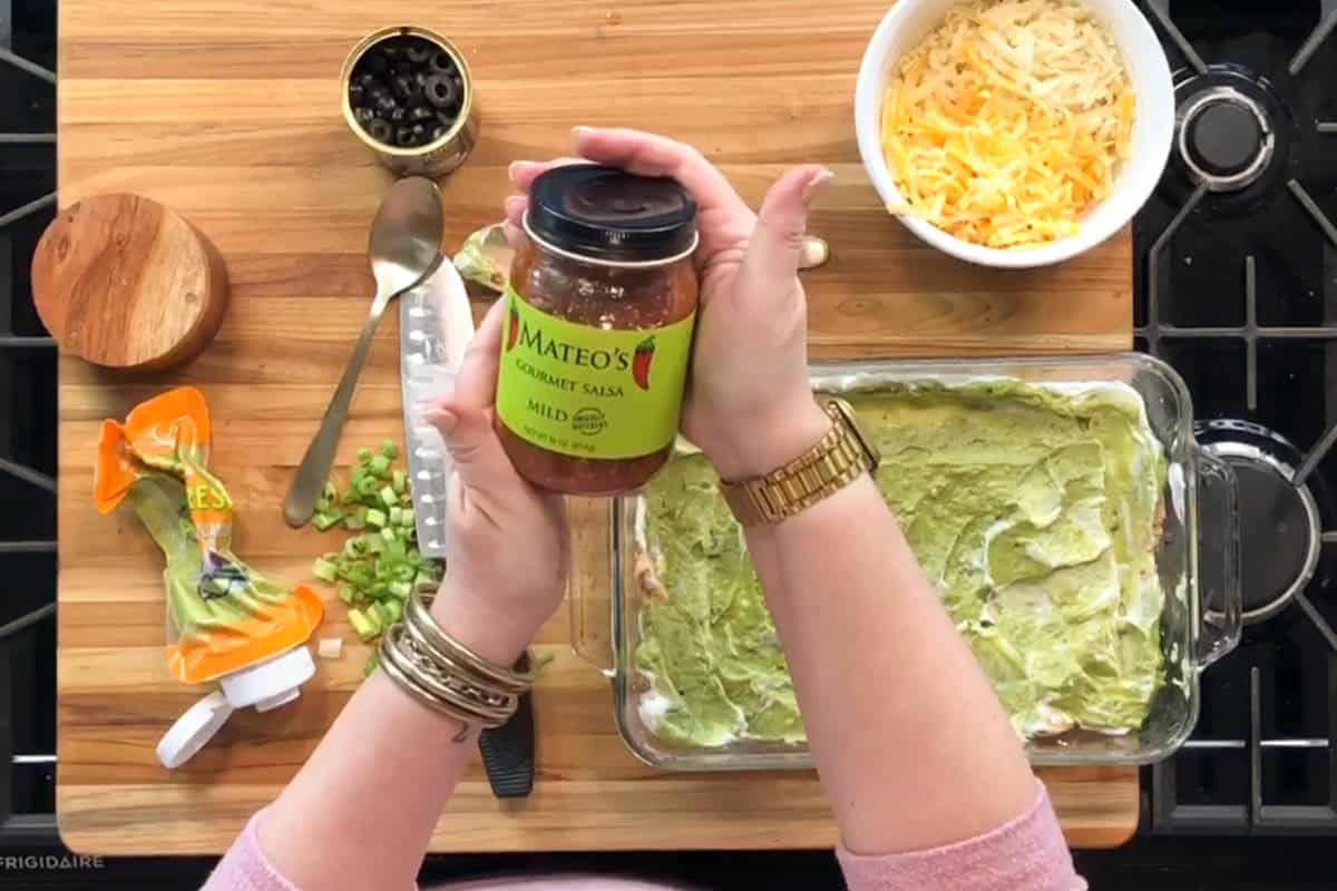 A person holds a jar of Mateo's Gourmet Salsa over a kitchen counter with a glass dish of guacamole, a bowl of shredded cheese, chopped green onions, sliced olives, a spoon, and other ingredients.