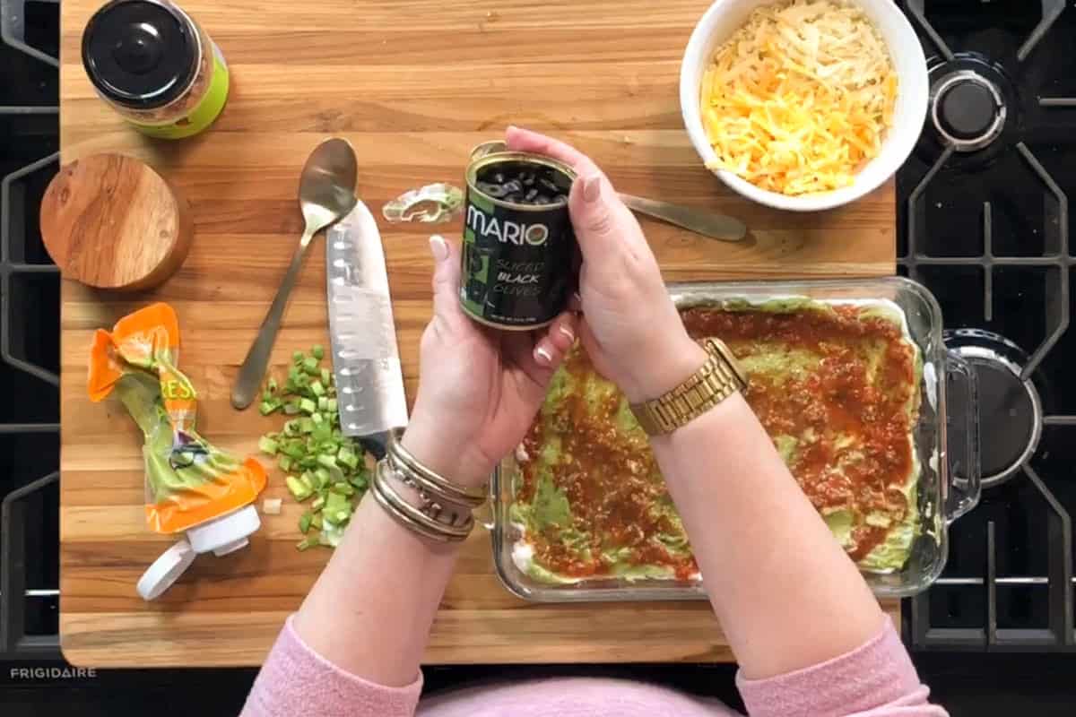 A person holds an open can of sliced black olives above a glass dish with layered dip on a wooden countertop. Nearby are chopped green onions, shredded cheese, a spoon, a knife, and an olive jar.