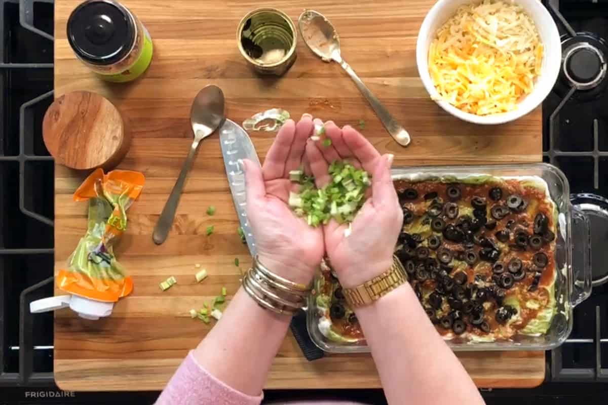 A person holding chopped green onions over a casserole topped with sliced black olives, next to a bowl of shredded cheese, utensils, an empty can, and other ingredients on a wooden cutting board.