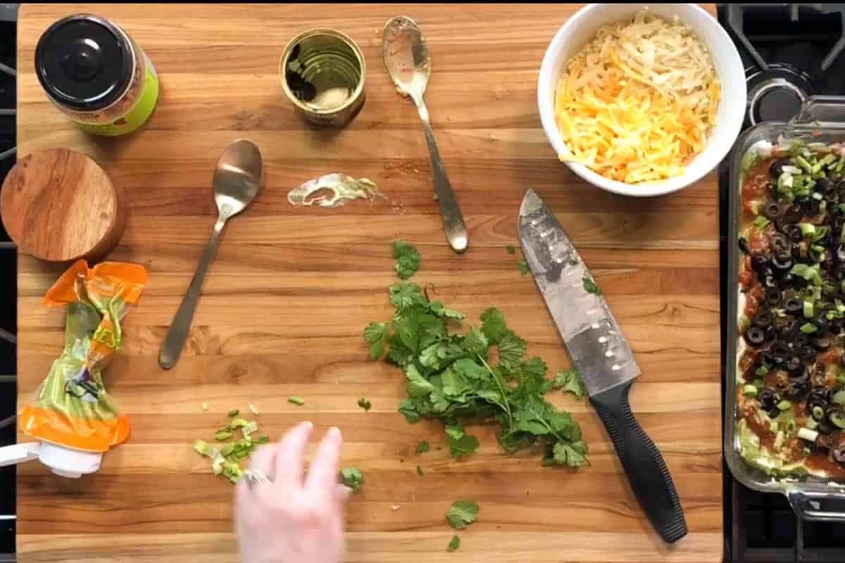 A wooden cutting board with chopped cilantro, a partially sliced bundle of herbs, two spoons, a knife, an open can, a jar, shredded cheese in a bowl, and a hand reaching for cilantro. A baking dish with food is at the edge.