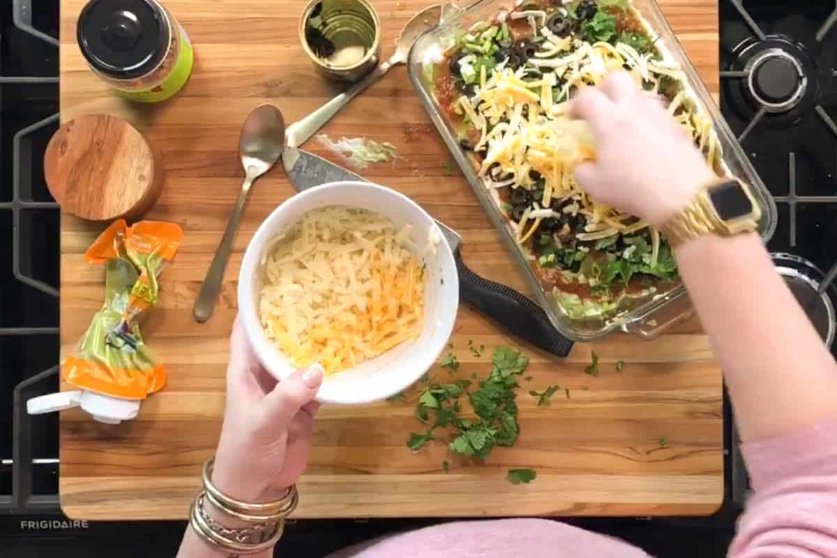 A person sprinkles shredded cheese over a casserole in a glass dish. There is a bowl of cheese, a packet of cheese, fresh cilantro, a can, and a wooden container on a wooden cutting board next to a stove.