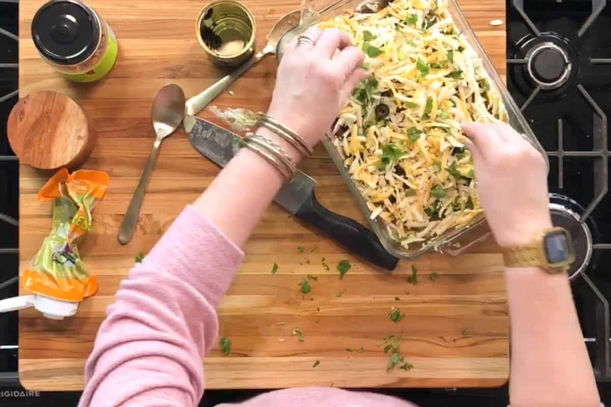 A person wearing a pink top and gold watch sprinkles shredded cheese and herbs onto a casserole in a glass baking dish. Various kitchen utensils and ingredients are on the wooden counter.