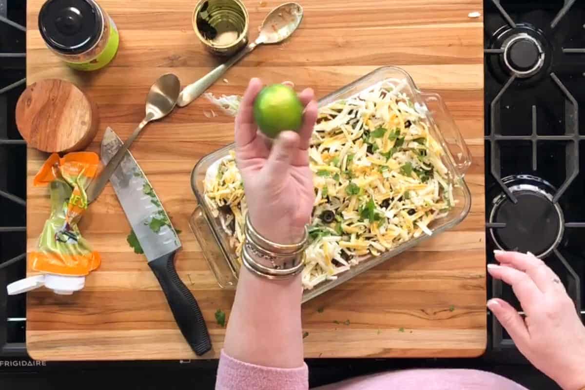 A person holds a lime above a glass baking dish filled with shredded cheese and herbs on a wooden cutting board, with a knife, spoons, jar, and cheese packaging nearby.
