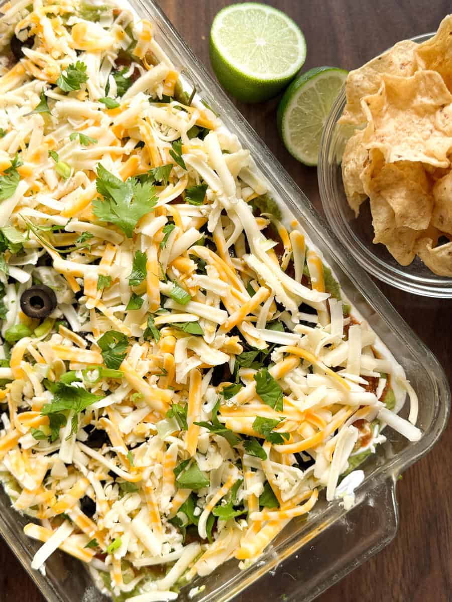 A glass dish filled with layered dip topped with shredded cheese, sliced olives, and cilantro sits next to a bowl of tortilla chips and a halved lime on a wooden surface.