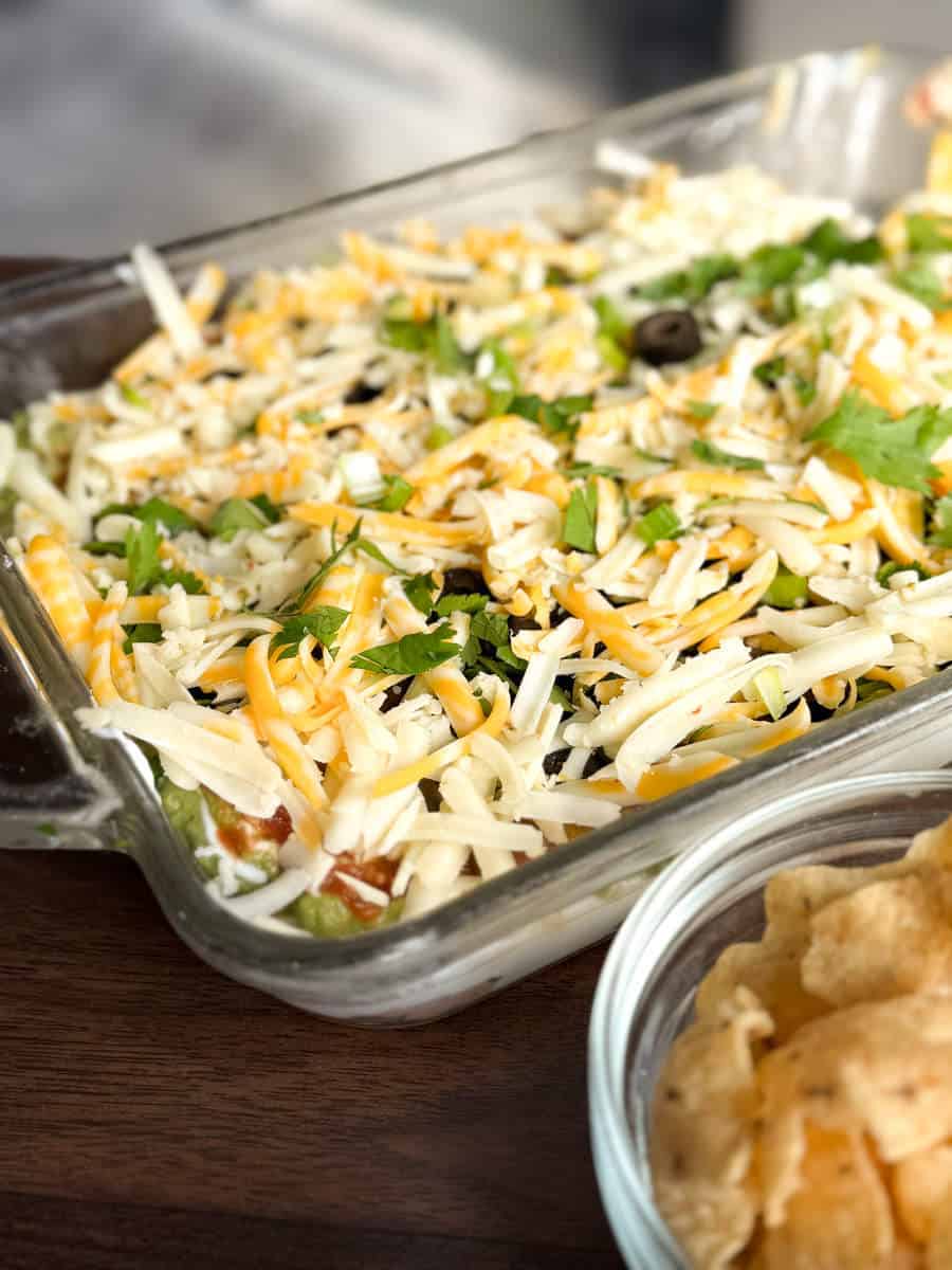 A glass baking dish filled with a layered dip topped with shredded cheese, chopped cilantro, and black olives sits on a wooden surface next to a bowl of tortilla chips.