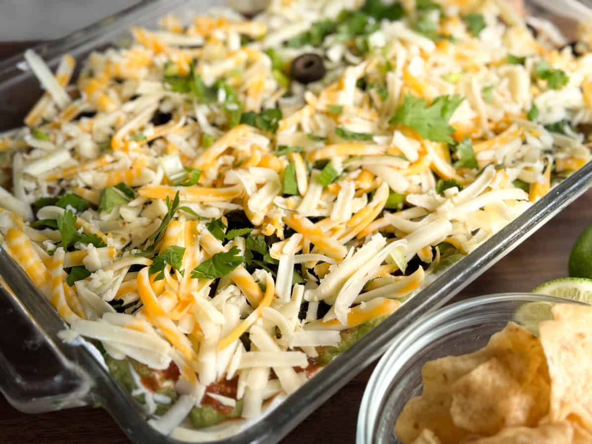 A glass baking dish filled with layered dip topped with shredded cheese, sliced olives, and fresh cilantro. A bowl of tortilla chips and lime wedges are visible in the foreground.