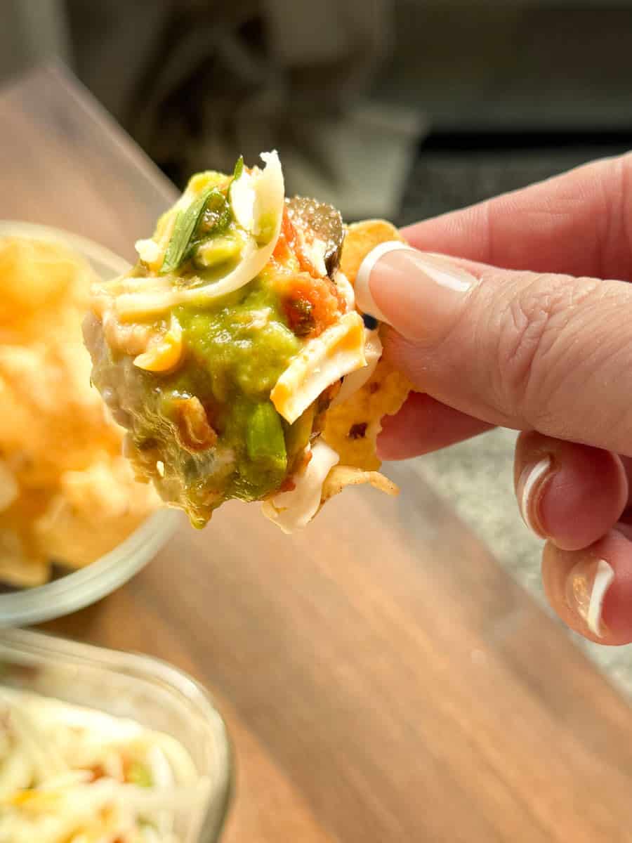 A hand holding a tortilla chip topped with shredded cheese, guacamole, salsa, and other toppings. A bowl of chips and another dish are visible in the background on a wooden surface.