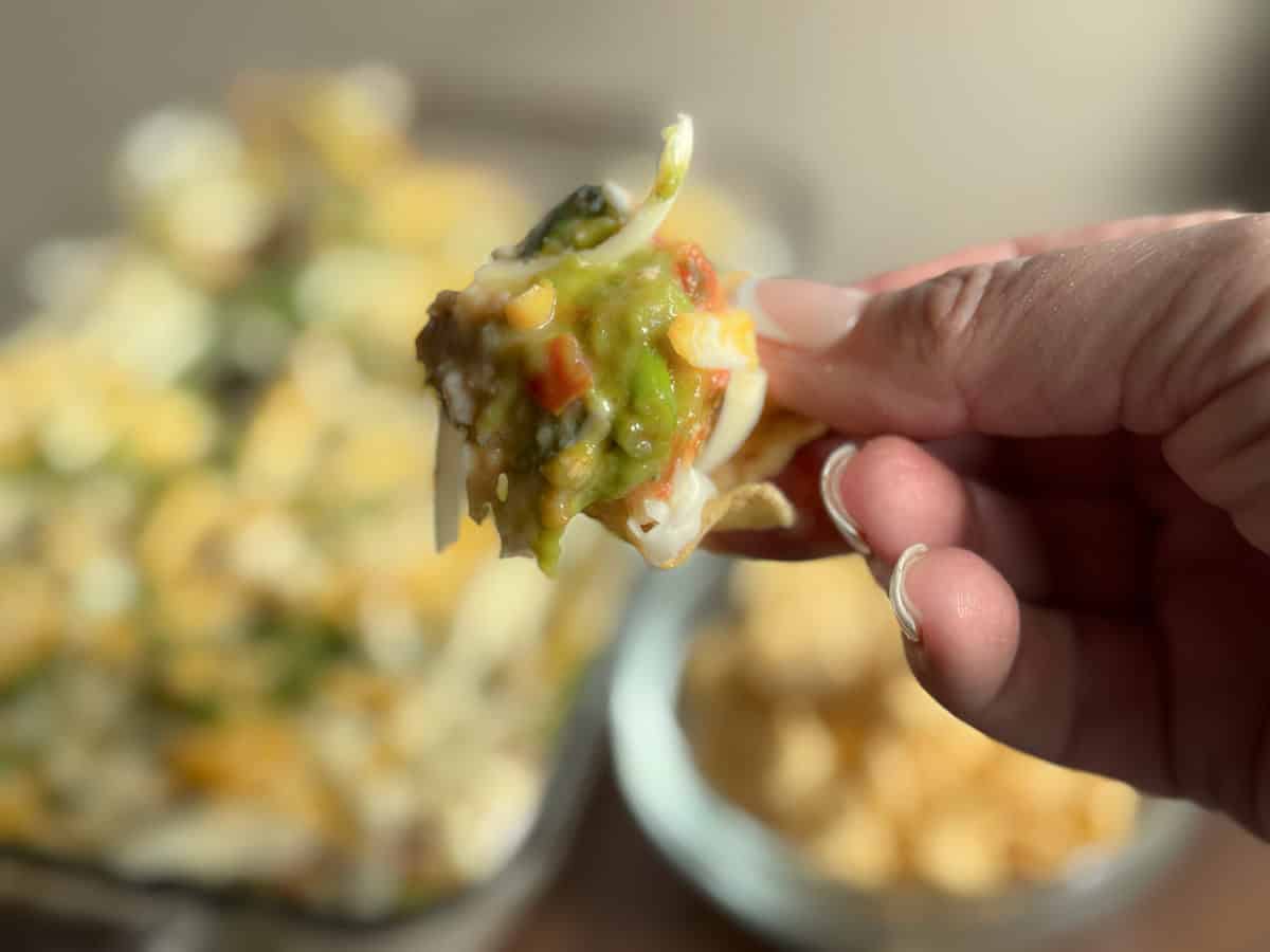 A hand holds a corn chip topped with layers of guacamole, cheese, and other dip ingredients. A glass dish with more layered dip and a bowl of corn chips are seen blurred in the background.