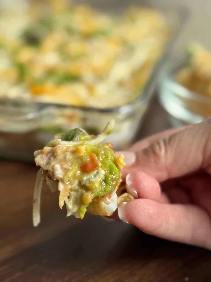 A close-up of a hand holding a chip topped with layered dip containing beans, cheese, guacamole, and chopped vegetables, with a glass dish of the dip blurred in the background.
