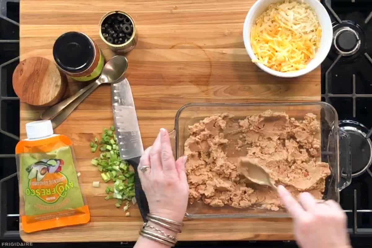 A person spreads refried beans in a glass dish on a wooden cutting board. Nearby are chopped green onions, a packet of guacamole, measuring spoons, a can of sliced olives, a jar, and a bowl of shredded cheese.