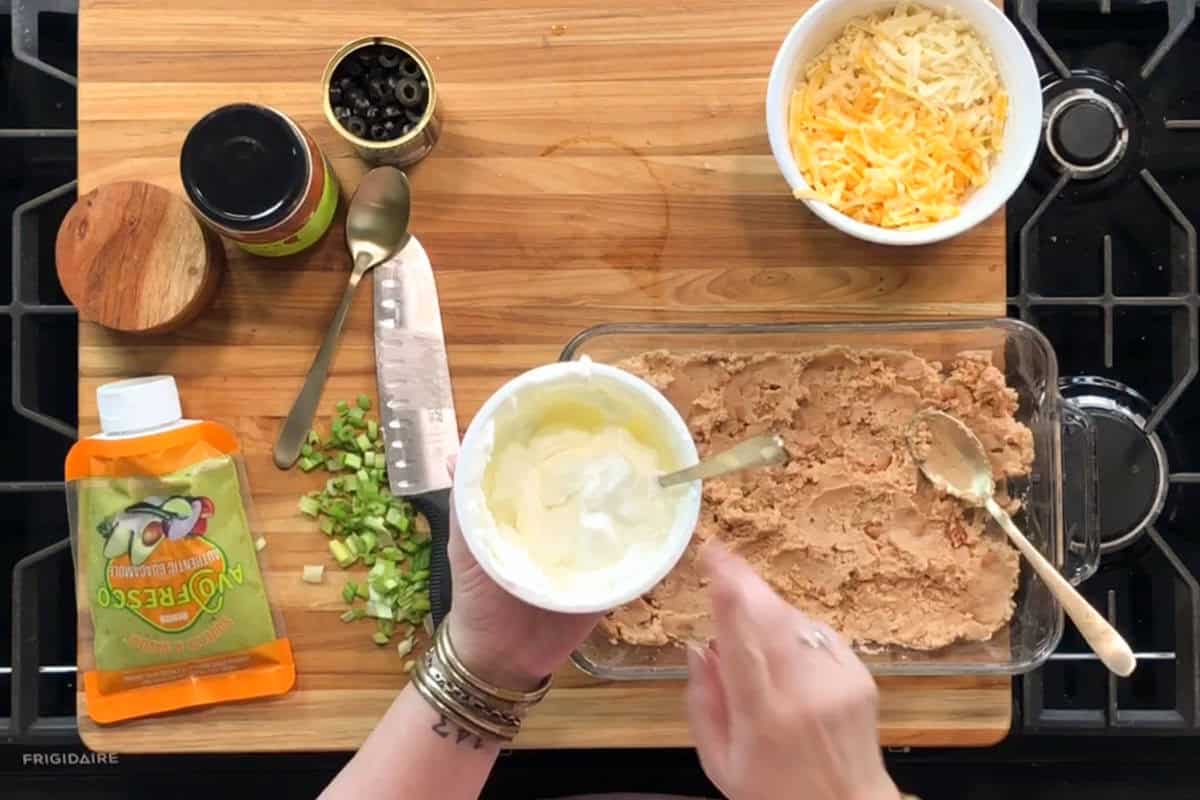 A person spreads sour cream into a glass dish of refried beans. Other ingredients, such as shredded cheese, chopped green onions, sliced black olives, and guacamole, are arranged on a wooden cutting board on a stovetop.