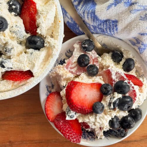 A bowl and a plate filled with a dessert made of whipped cream, sliced strawberries, and blueberries, with a spoon resting in the plate and a blue and white cloth nearby on a wooden surface.
