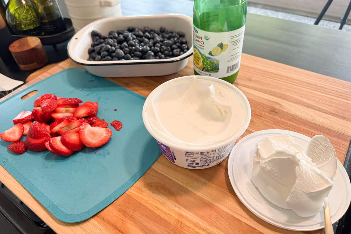 A blue cutting board with sliced strawberries, a tray of blueberries, a bottle of lemon juice, a tub of whipped topping, and a block of cream cheese sit on a wooden countertop.