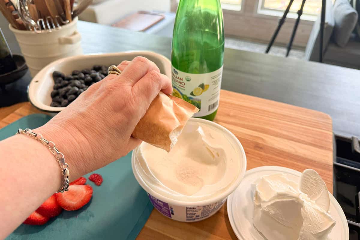 A hand pours contents from a brown packet into a container of white creamy substance on a wooden counter. Sliced strawberries, blueberries, and a green bottle are nearby.