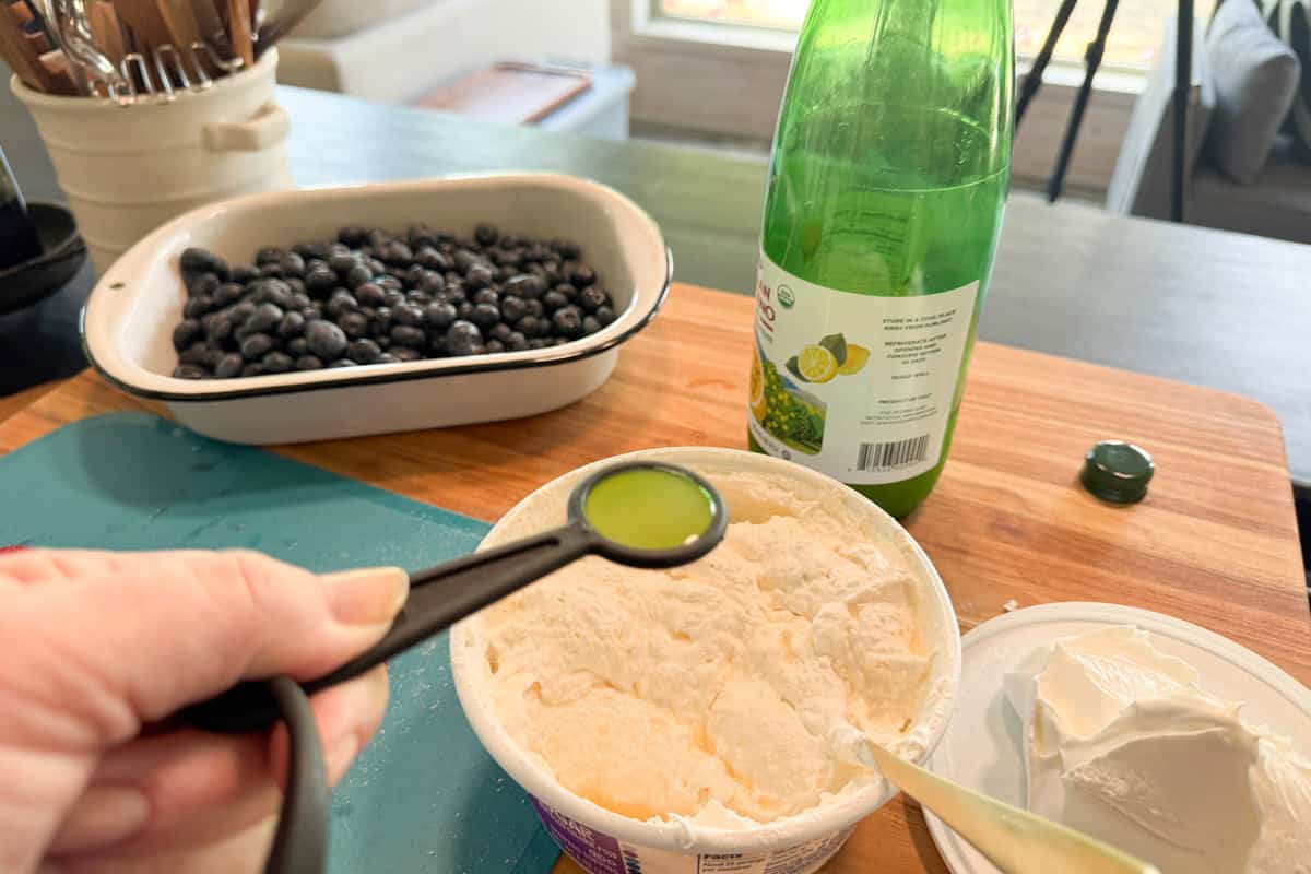 A hand holds a tablespoon of lime juice over a container of cottage cheese on a wooden counter. Nearby are a dish of blueberries, a lime juice bottle, and a block of cream cheese.