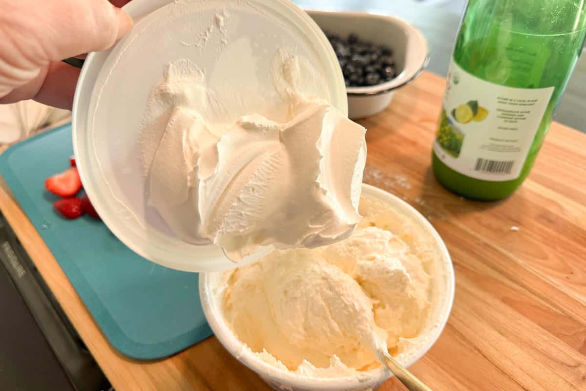 A hand holds a white plastic lid with whipped cream on it above an open container of whipped cream on a wooden countertop. Nearby are a bottle of lime juice, a bowl of blueberries, and sliced strawberries on a cutting board.