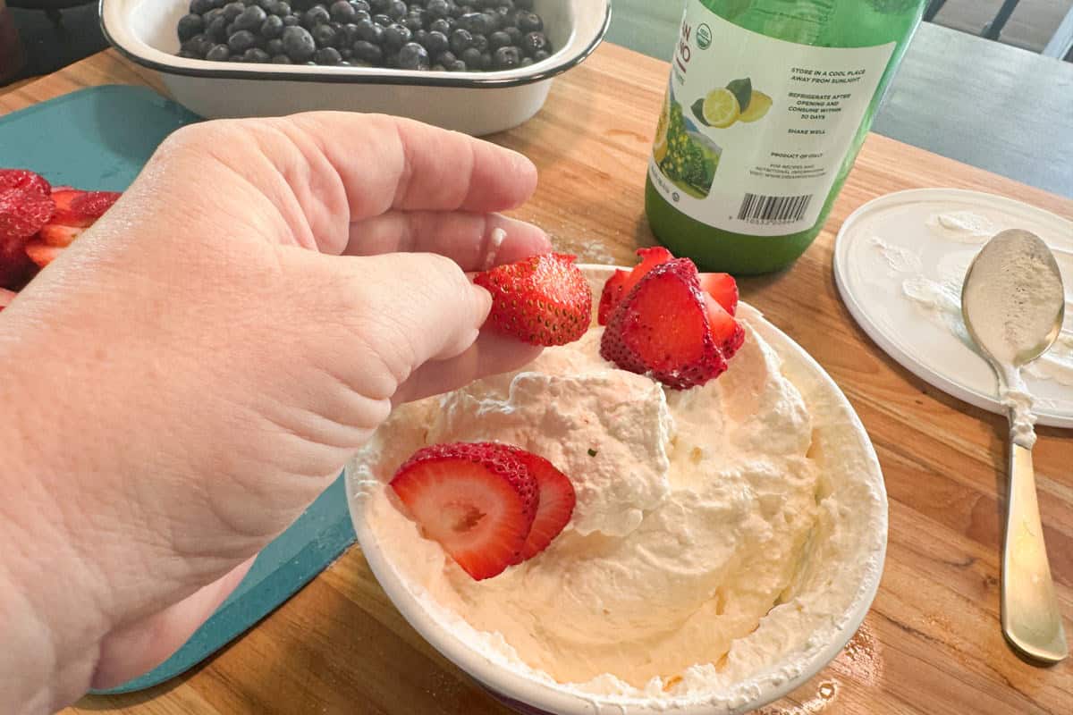 A hand holds a strawberry half and dips it into a bowl of whipped cream. Sliced strawberries, a bowl of blueberries, a green bottle, and a spoon on a plate are on the wooden surface nearby.